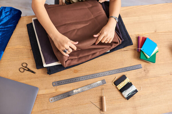 cropped view of dressmaker near quality fabrics samples and sewing supplies on work desk in atelier
