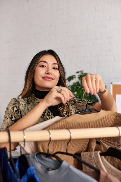 привлекательный счастливый азиатский стилист с модной одеждой на заказ, смотрящий в камеру возле стойки в собственной студии