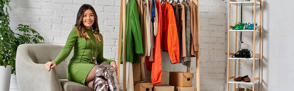 веселый стильный азиатский дизайнер в кресле возле стойки со специальной одеждой в ателье моды, баннер