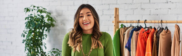 творческий успешный азиатский дизайнер, смотрящий на камеру возле стойки с одеждой в собственном ателье, баннер