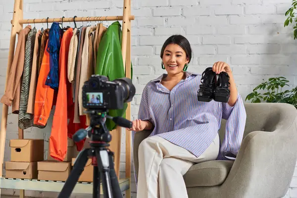 stock image pleased asian clothes designer showing trendy footwear during video blog in personal fashion studio