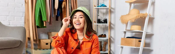 stock image joyful asian stylist posing in orange attire and panama hat in atelier, fashion business, banner