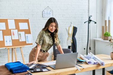 young smiling asian clothes designer looking at laptop on work desk in personal fashion atelier clipart