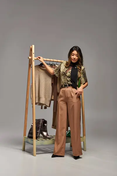 时尚的亚洲服装设计师站在靠近衣架的地方 穿着灰色背景的时髦定制服装 — 图库照片