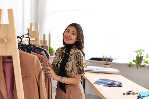 Neşeli Asyalı moda tasarımcısı özel atölyede özel dikim kıyafetlerle kameraya bakıyor.