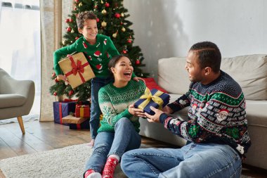 Neşeli Afrikalı Amerikalı kadın, yanlarında oğulları varken kocasından Noel hediyesi alıyor.