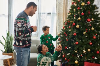 Neşeli Afro-Amerikan ailesi Noel ağacını mücevherlerle süslüyor ve mutlu bir şekilde gülümsüyor.