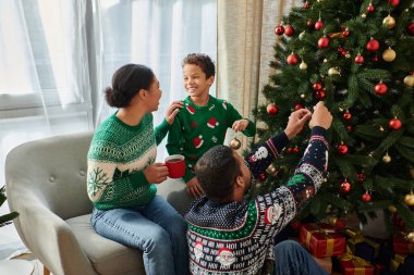 Neşeli Afrikalı Amerikalı kadın, oğlu ve kocası Noel ağacı süslerken kahve içiyor.