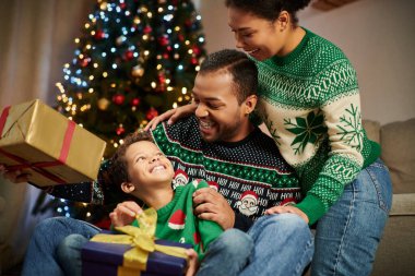 Neşeli, tapılası Afrikalı Amerikalı çocuk arka planda bulanık Noel ağacıyla ailesine gülümsüyor.
