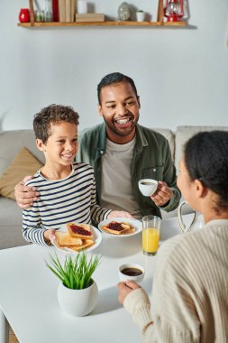 vertical focused shot of happy african american father and son hugging and looking at blurred mother clipart