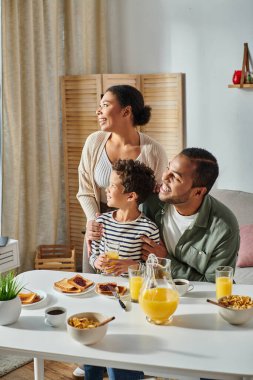 vertical shot of happy modern african american family in homewear looking away during breakfast clipart