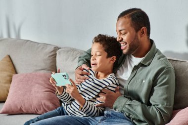 Mutlu Afro-Amerikan baba ve oğlu rahat kıyafetler içinde koltukta oturmuş cep telefonlarına bakıyorlar.
