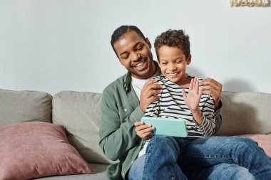 cheerful african american boy sitting on his father laps and waving at mobile phone camera clipart