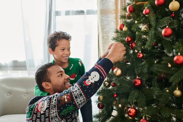 stock image happy african american father and son wearing cozy sweaters decorating Christmas tree with baubles