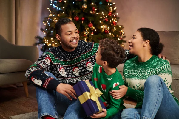 joyous african american man looking happily at his son and wife sitting on floor by Christmas tree