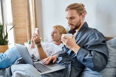 bearded gay man with coffee cup networking on laptop near tattooed boyfriend with smartphone on bed clipart