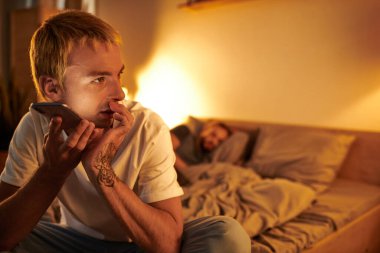 Dövmeli sadakatsiz eşcinsel adam akıllı telefondan sesli mesaj yolluyor. Yatak odasında uyuyan erkek arkadaşın yanında.