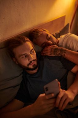 sadakatsiz sakallı eşcinsel adam gece yatak odasında uyuyan erkek arkadaşın yanında akıllı telefondan sohbet ediyor.