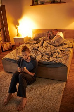 unfaithful bearded gay man messaging on mobile phone near sleeping boyfriend at night in bedroom clipart