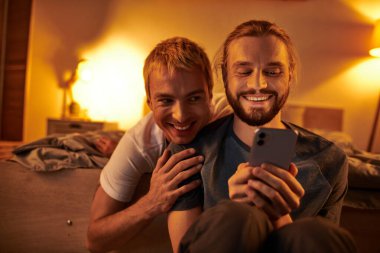 joyful bearded gay man browsing internet on mobile phone near smiling boyfriend in bedroom at night clipart