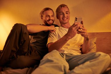 smiling gay man browsing social media on smartphone near happy boyfriend in bedroom at night clipart