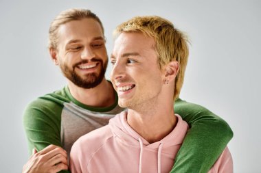 joyful bearded gay man embracing stylish boyfriend smiling and looking away on grey backdrop clipart