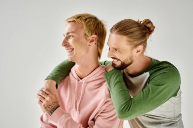 cheerful and stylish gay couple embracing and looking away while standing on grey backdrop clipart