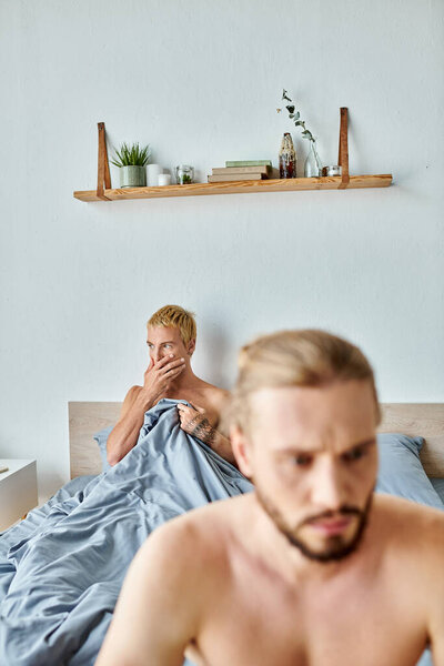worried gay man covering mouth with hand near offended boyfriend on blurred foreground in bedroom