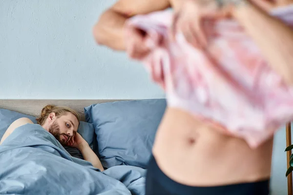 stock image upset bearded gay man lying on bed near love partner dressing up in bedroom, relationship issues