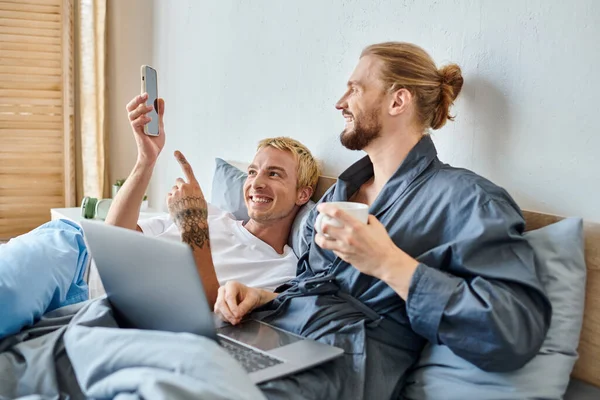 gay man with coffee cup smiling near laptop and tattooed boyfriend pointing at smartphone on bed