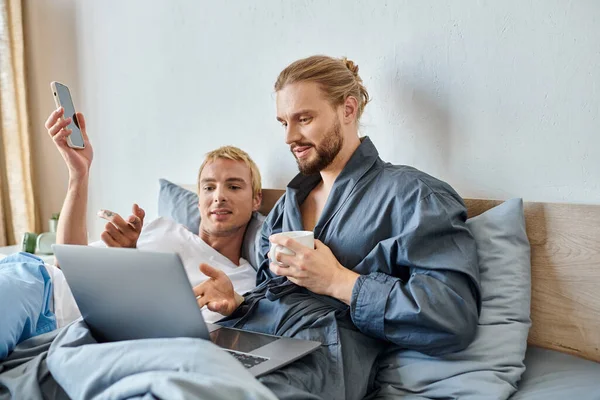 bearded gay man with coffee cup pointing at laptop near tattooed boyfriend with smartphone on bed