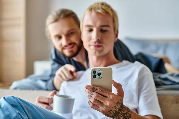 stock image tattooed gay man with coffee cup browsing internet on mobile phone near smiling boyfriend in bedroom