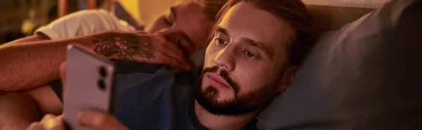 disloyal bearded gay man chatting on smartphone near sleeping boyfriend at night in bedroom, banner