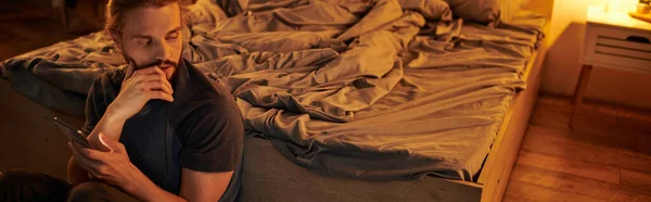 stock image bearded thoughtful man with mobile phone sitting near bed at high in bedroom, horizontal banner