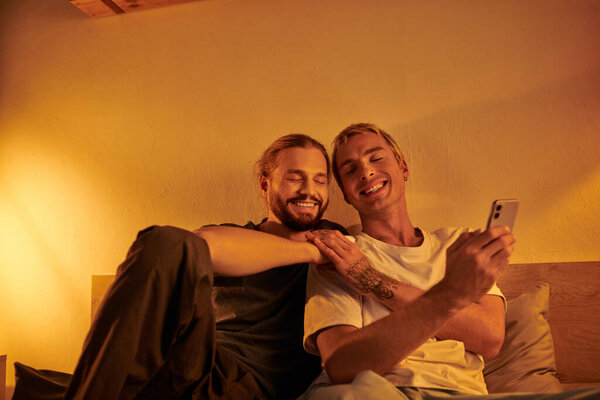 happy tattooed gay man holding smartphone near smiling bearded boyfriend in bedroom at night