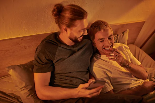 stock image bearded gay man browsing social media on smartphone near laughing boyfriend in bedroom at night