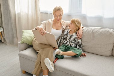 mother reading book to her joyful daughter with prosthetic leg and sitting together in living room clipart