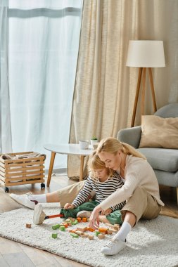 blonde girl with prosthetic leg sitting on carpet and playing wooden blocks game near mother clipart