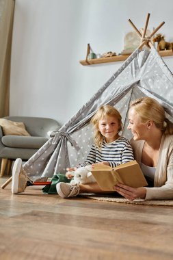 Protez bacaklı mutlu kız annesinin yanında oyuncak tutuyor evde oyun çadırında kitap okuyor.