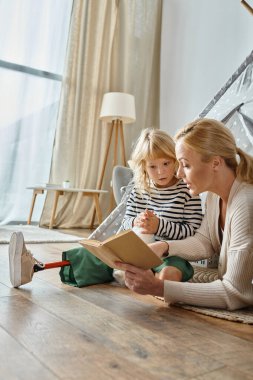Protez bacaklı tatlı bir kız yumuşak bir oyuncak tutuyor ve annesinin oyun çadırında kitap okumasını dinliyor.