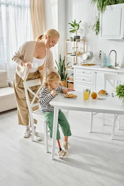 Stock image happy woman standing near daughter with prosthetic leg having breakfast in kitchen, inclusion