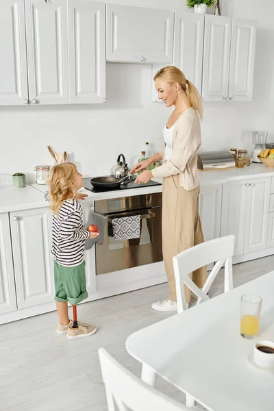 キッチンで料理を洗う幸せな母親を助ける義足の保持プレートを持つかわいい少女 — ストック写真