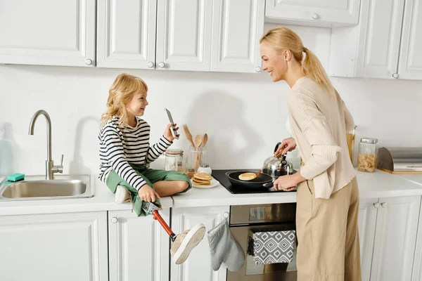 Protez bacaklı kız tezgahta oturuyor ve annesine krep pişiren akıllı telefonu gösteriyor.