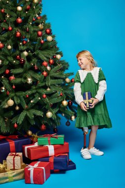 Protez bacaklı neşeli küçük kız hediyesini mavi üzerine sarmış Noel ağacının yanında