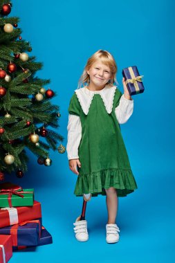 Protez bacaklı mutlu küçük kız hediyesini mavi üzerinde Noel ağacının yanında tutuyor.