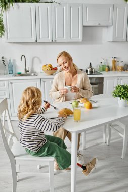 smiling blonde mother looking at her cute daughter with prosthetic leg having breakfast in kitchen clipart