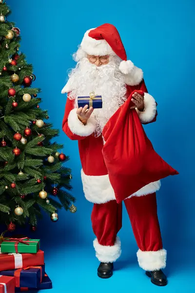 Sakallı ve gözlüklü mutlu Noel Baba çanta tutuyor ve Noel ağacının yanında duruyor.