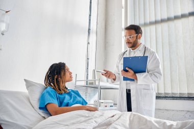 Genç Hintli doktor, Afro-Amerikan hastasına bakıyor. Hastane koğuşunda yatıyor.