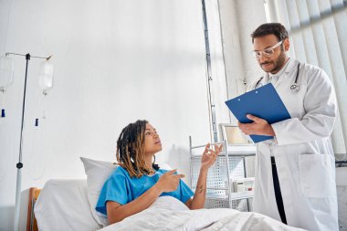 Genç Hintli doktor, Afro-Amerikan hastasına bakıyor. Hastane koğuşunda yatıyor.
