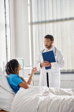 handsome indian doctor in glasses talking to young african american patient in hospital, healthcare clipart